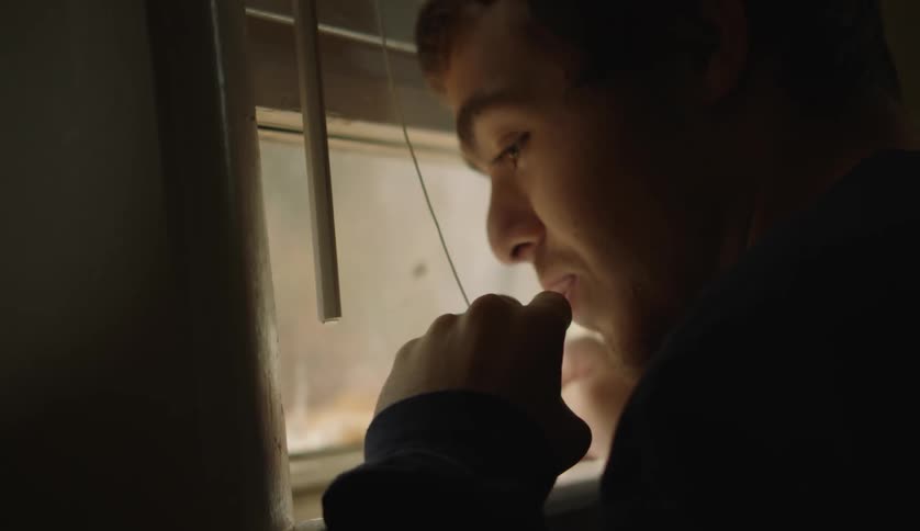 A teenage boy is standing in a room looking out the window