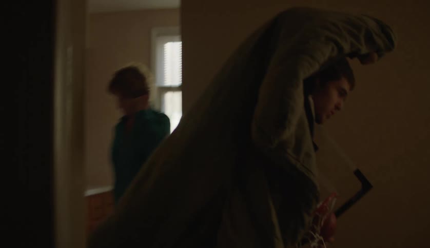 A teenage boy walks through the house and into a room carrying a blanket and a basketball hoop