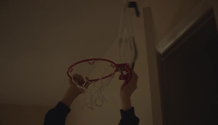 A young man hangs a miniature basketball hoop from a door in a house during the day