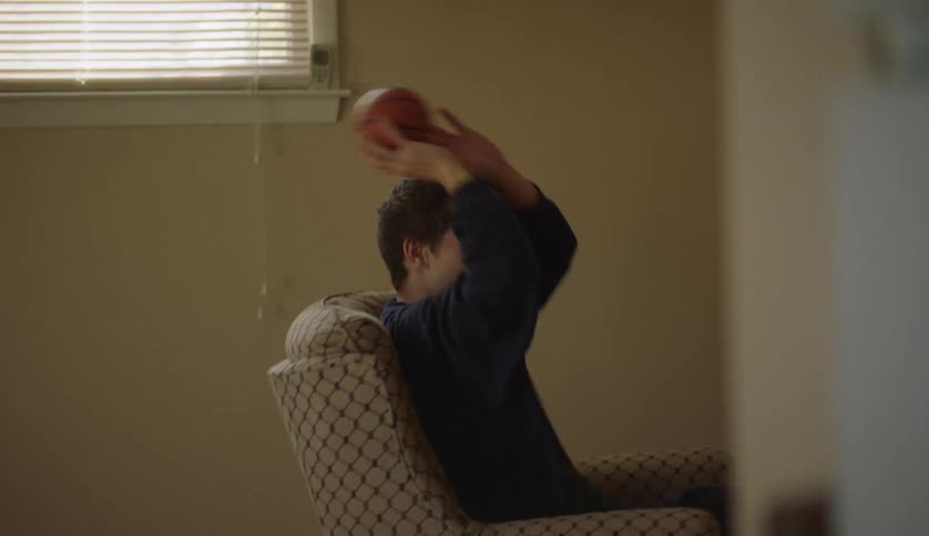 A teenage boy is sitting in a living room chair holding a ball as he raises his arm and tosses the ball