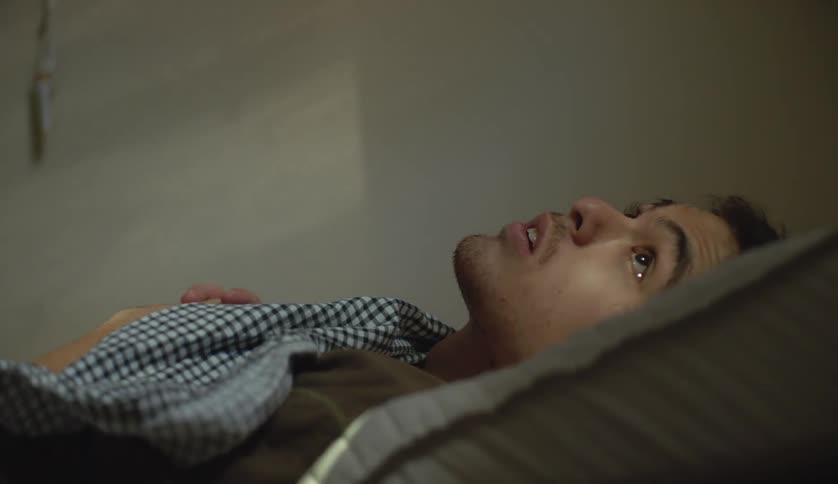 A teenage boy is lying in bed sleeping and he looks up when something wakes him