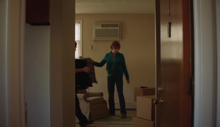 A woman moves a pillow on to a chair at home and her son carries in a television and sets it down on a stand