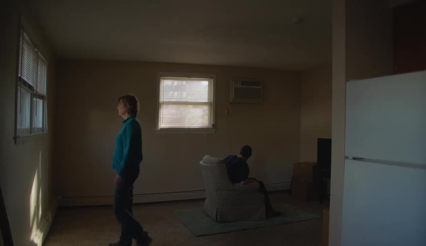 Older woman walks to and looks out window as young man sits in front of television in lightly furnished room