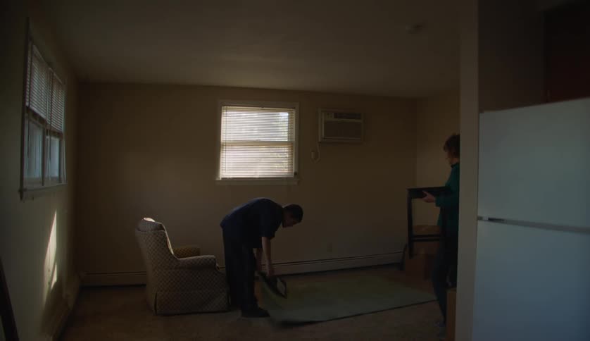 A teenage boy carries a rug into a room and lays it out on the floor as his mother brings a small table