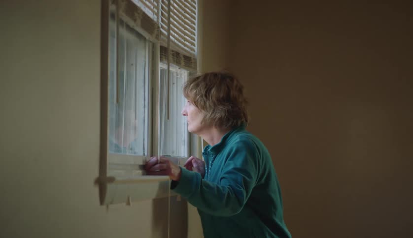 Middle aged woman with blue shirt and jeans looks out the window in room