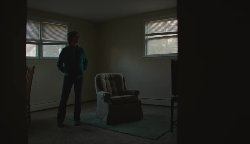 Middle aged woman wearing light blue jacket and jeans stands next to armchair in dim lit room