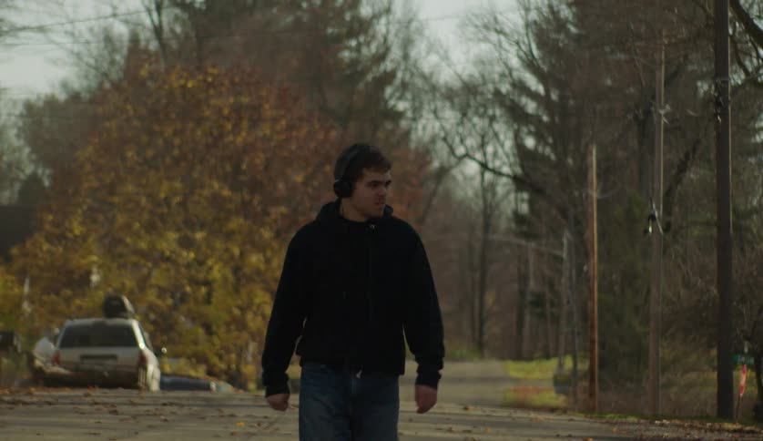 A teenage boy wearing headphones is walking on a neighborhood street lined with trees