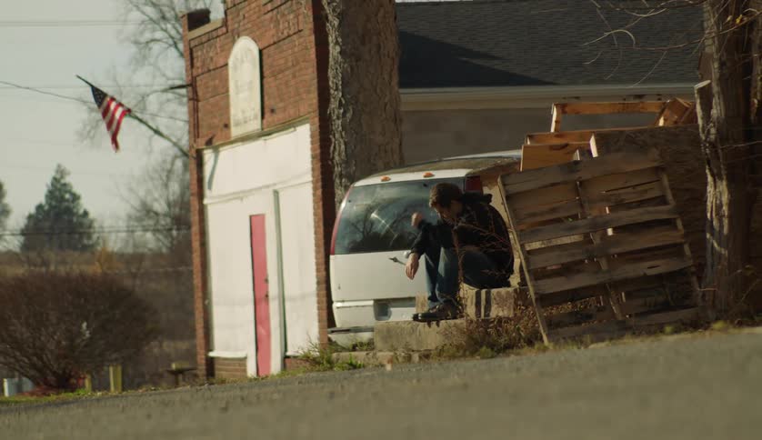 A teenage boy sits outside a home with his head down and scratches his hair
