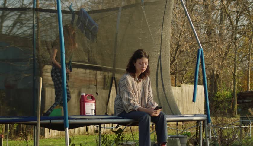 Teenager checks her phone sitting on a trampoline in the backyard