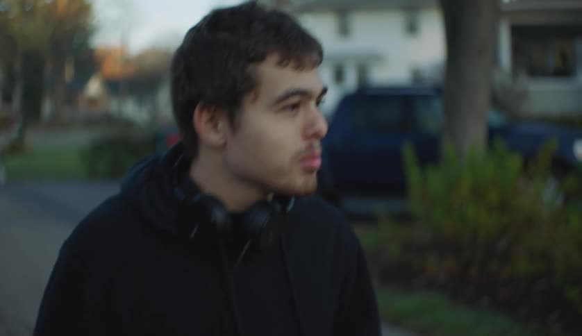 A teenage boy with headphones around his neck is walking on a neighborhood street and looking around