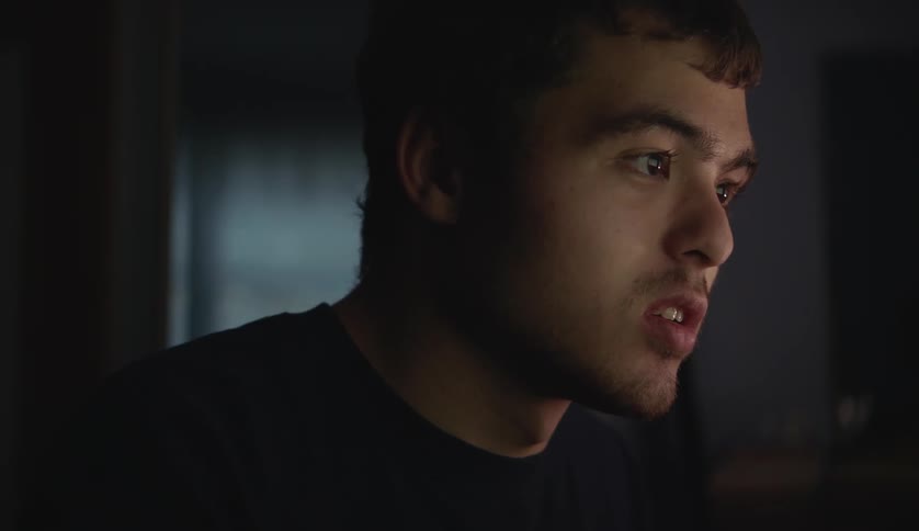 Light shines on the face of a teenage boy as he looks down at something then looks up and points then rocks back and forth