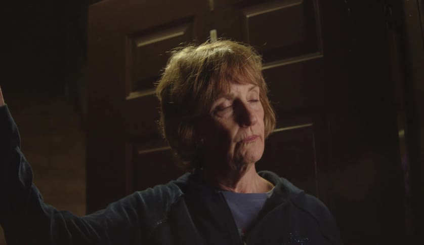Middle aged woman with brown hair and zip up hoodie opens door and talks to person