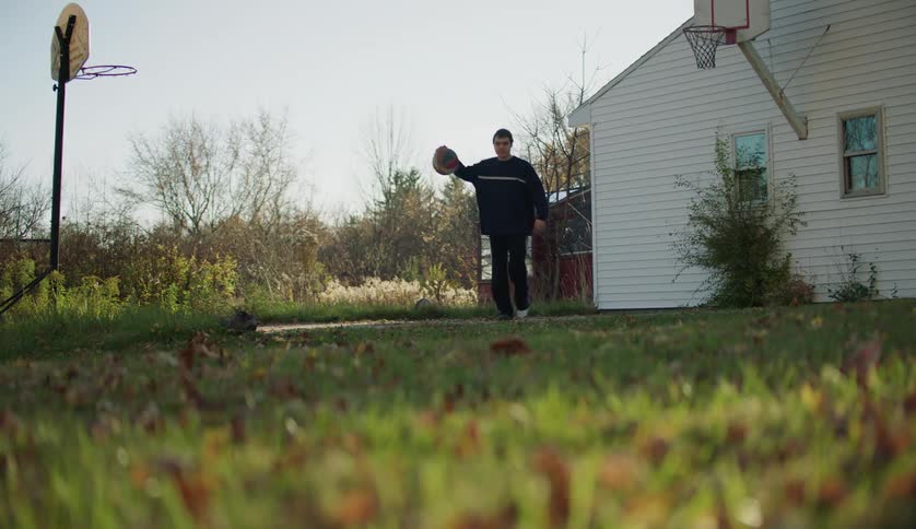 A teenage boy is outside of his house dribbling a basketball and misses a shot in the hoop