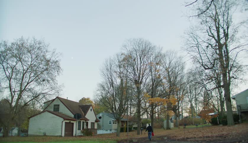 A teenage boy is walking on a neighborhood street carrying a red shirt as he walks to his house and opens the door
