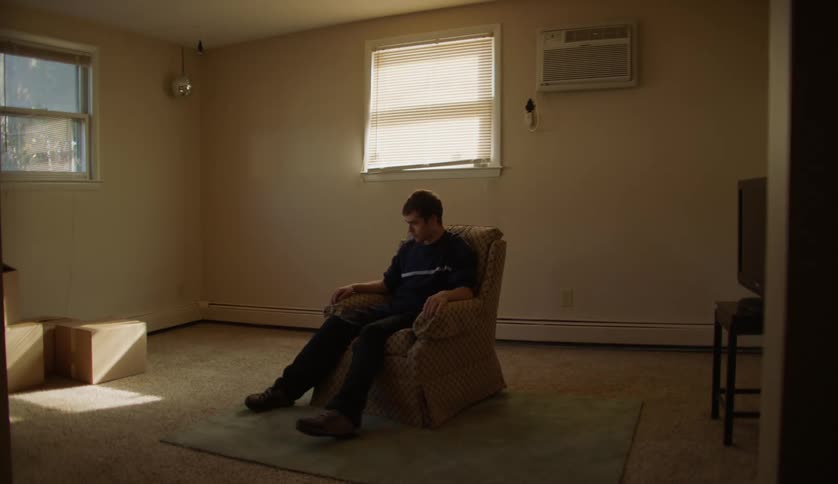 A young man swivels in an armchair in the mostly empty living room of a house during the day