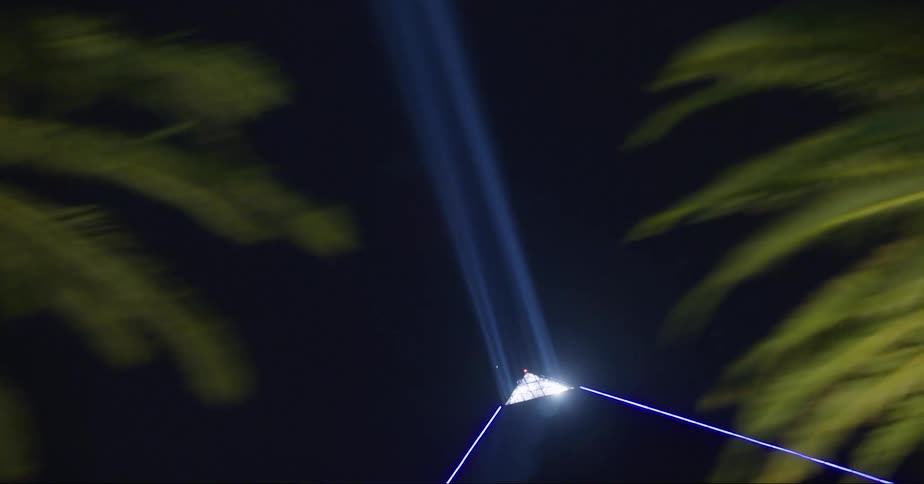 Palm tree lined las vegas street passes pyramid and sphinx replica as blue light shines from peak of pyramid at night