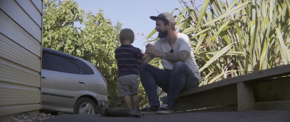 A young toddler boy plays with a small tabby cat on a sunny driveway.