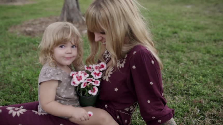 A little girl holding a pot of flowers as she bounces on a woman's lap and smiles