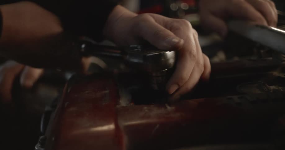 A man uses a wrench to loosen something under a hood of a car as another man watches