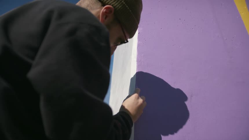 The sun shines upon a man as he stands outside painting a design on the wall of a building