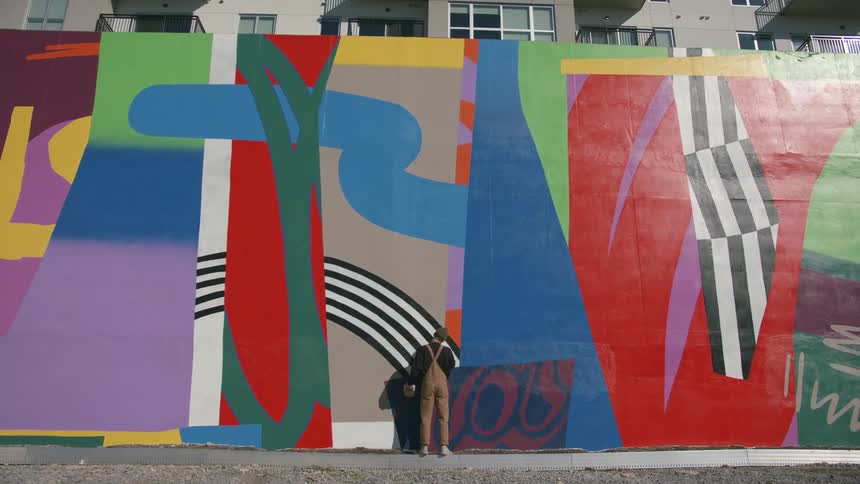 Man wearing brown overalls stands in front of a wall with an abstract mural