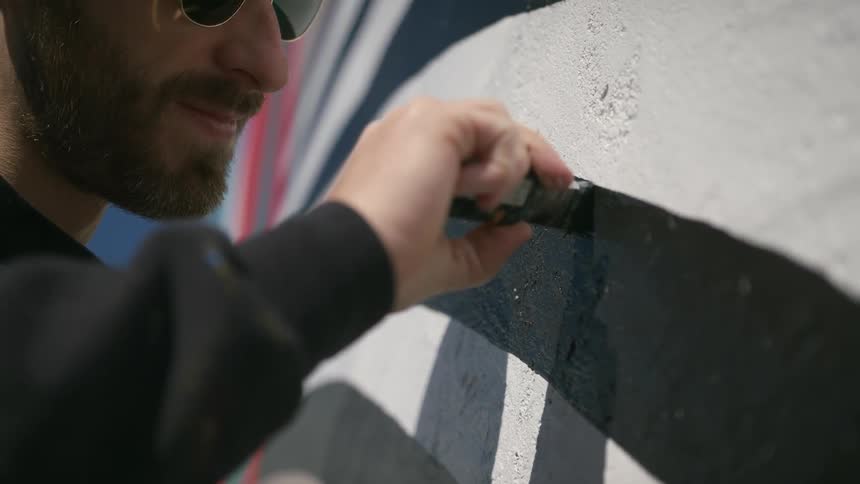 Man smiles while applying black paint to mural on stucco