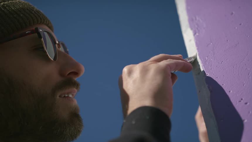 A man wearing sunglasses and a beanie paints a mural outdoors