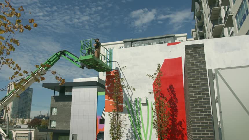 Man is using a lift truck to paint a mural on the side of a building in a city during the day
