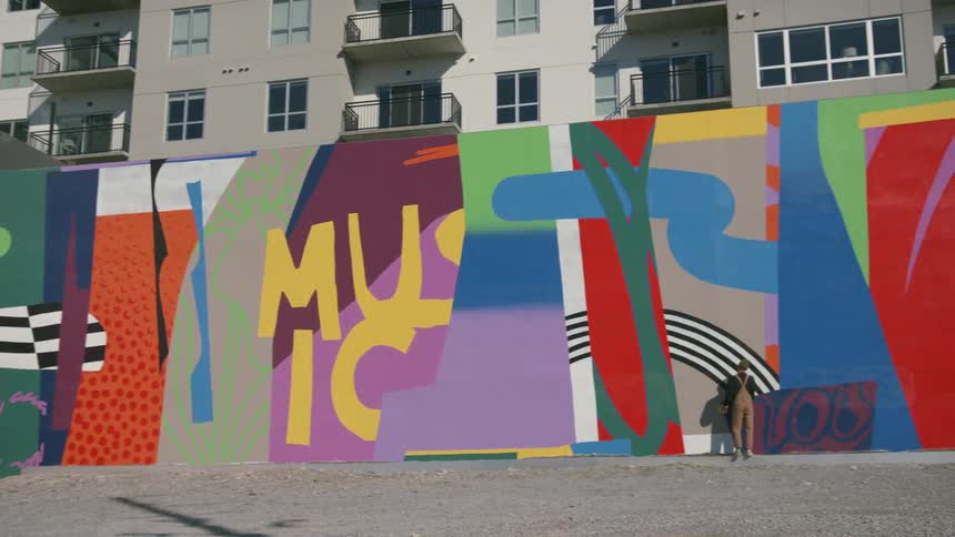 A painter stands in front of a wall with a large colorful mural in the city