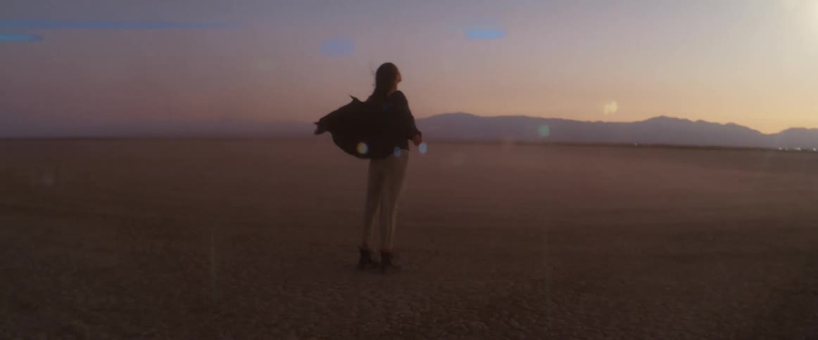 Woman in black shirt and gray pants stands and spreads her arms in the desert at dusk