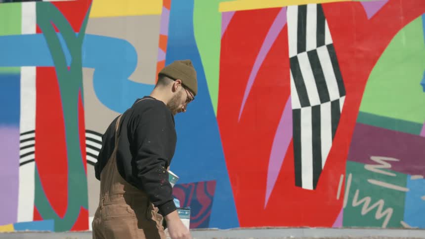 A man is carrying containers of paint as he walks near a wall with a colorful design painted on it