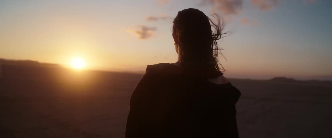 A woman is walking in the desert and the sun is shining brightly on the horizon and there are mountains in the distance