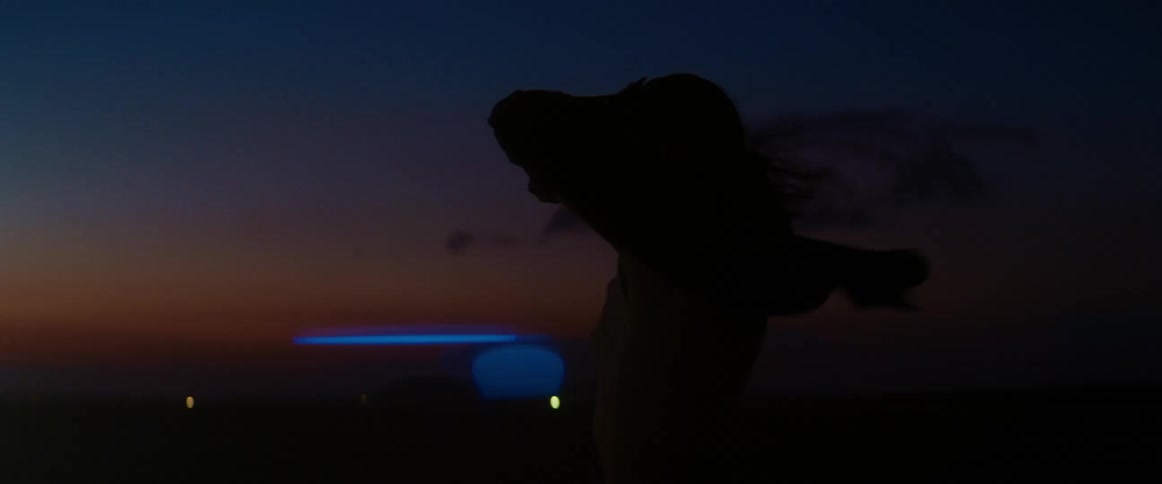 Wind blows womans hair as she raises her arm to block glaring light at night against a red horizon