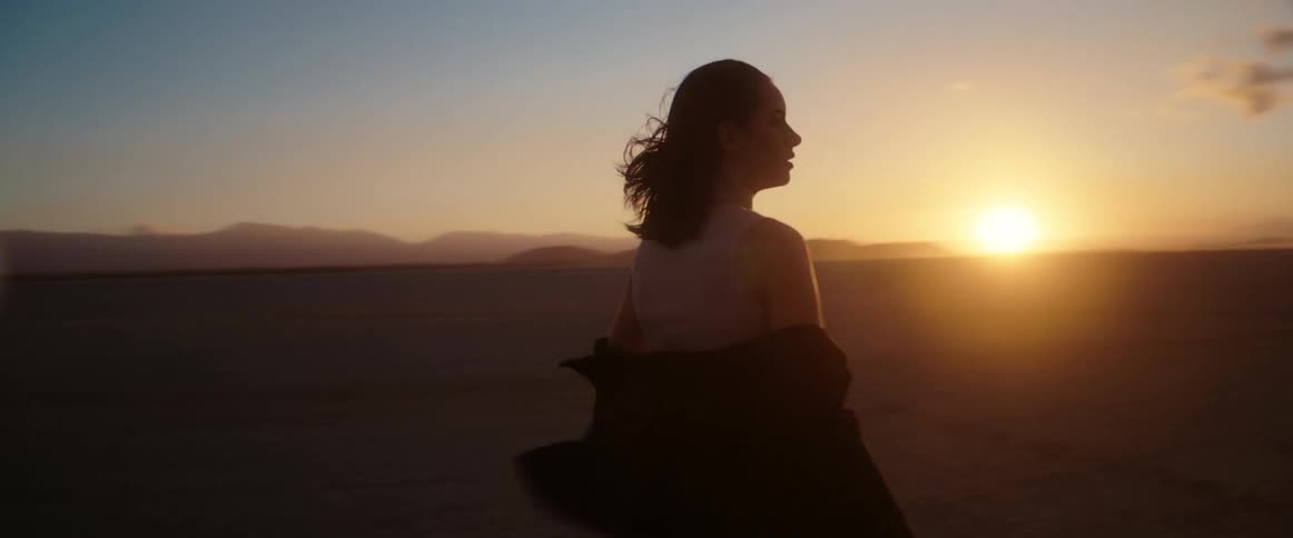 A young woman looks around at her surroundings while standing in a desert at sunset