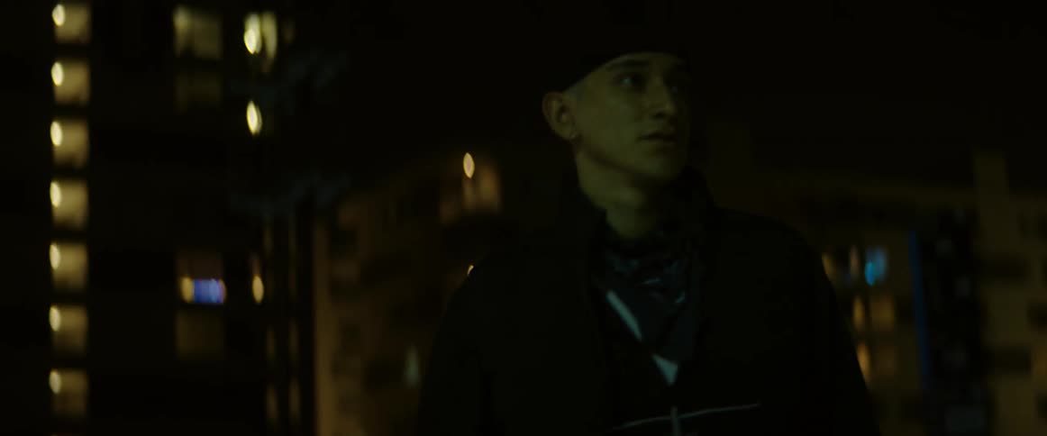 Man looks around city street at night as nearby spotlight creates a blue glare