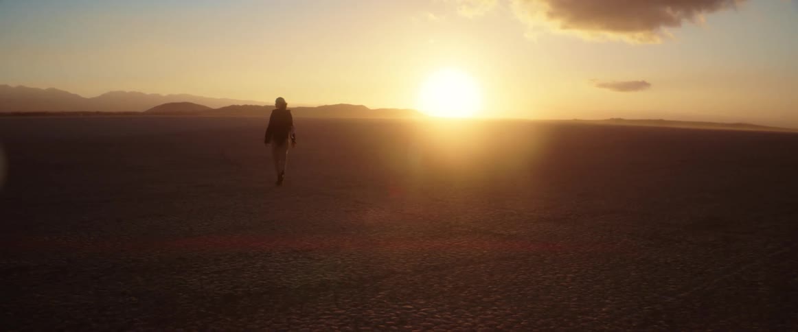 A woman is walking across the desert and the sun is shining brightly on the horizon and there are mountains in the distance