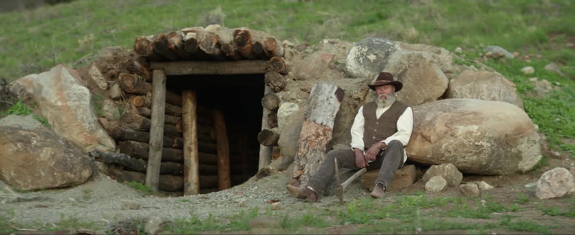 A man sitting outside a mine drives his pickaxe into the soil before staring into the camera