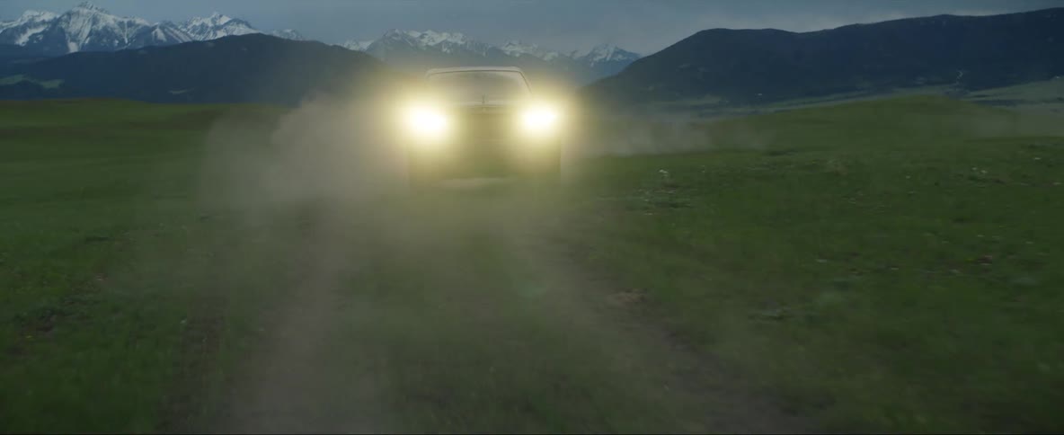 An automobile with the headlights on is traveling wildly and swerving on a rural road with mountains in the distance under a cloudy sky