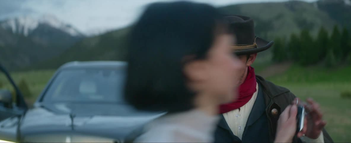 Man in a hat and red scarf dangles a key ring from his hand as a woman walks by while standing outdoors near a car