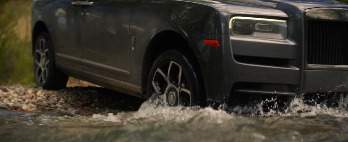 A vehicle drives through a shallow stream in a woodland during the day