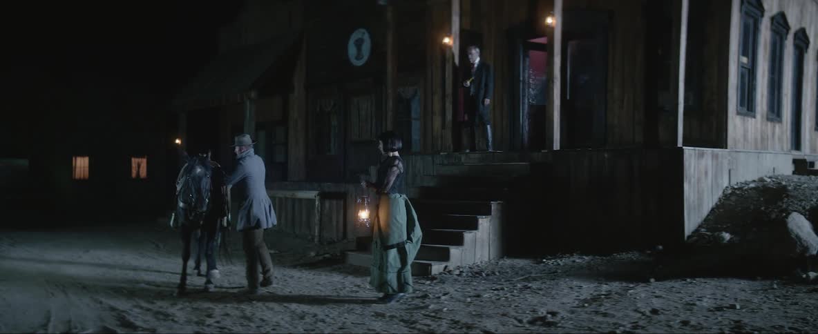 Woman holding a gas lamp waits while a man rides a horse out of a wild west town at night