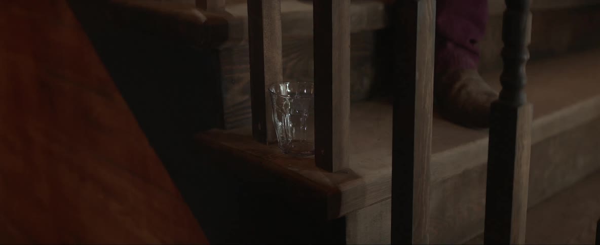 An empty glass is sitting at the edge of a step on a staircase as a man walks down the stairs kicking the glass off as the railing shakes