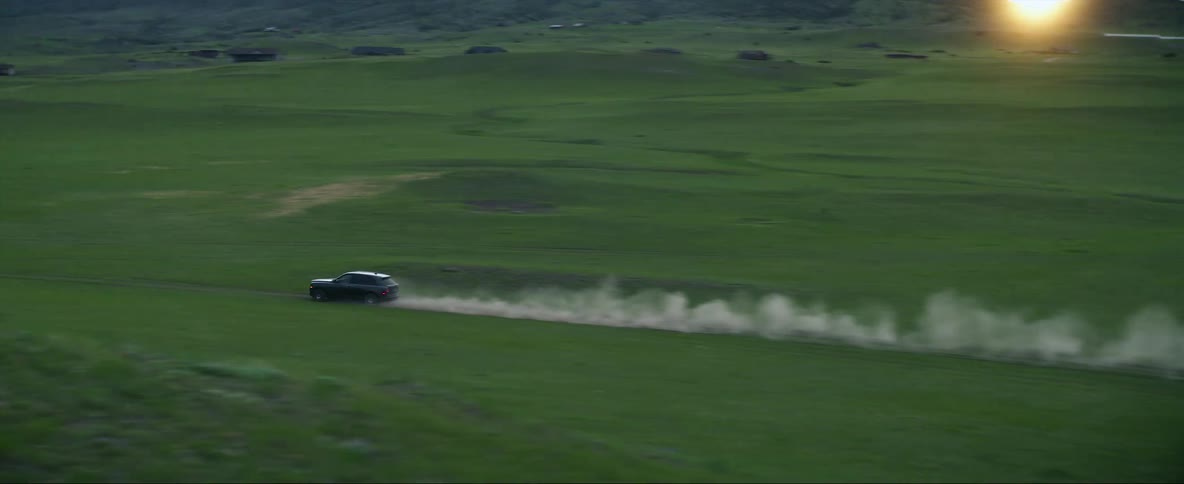 A vehicle sends up a cloud of dust behind it as it travels across a hilly grassland during the day