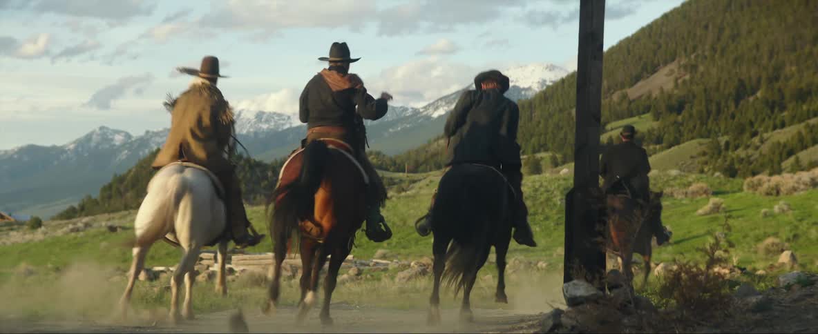 A group of men in old western attire ride off down a gravel road in the countryside during the day