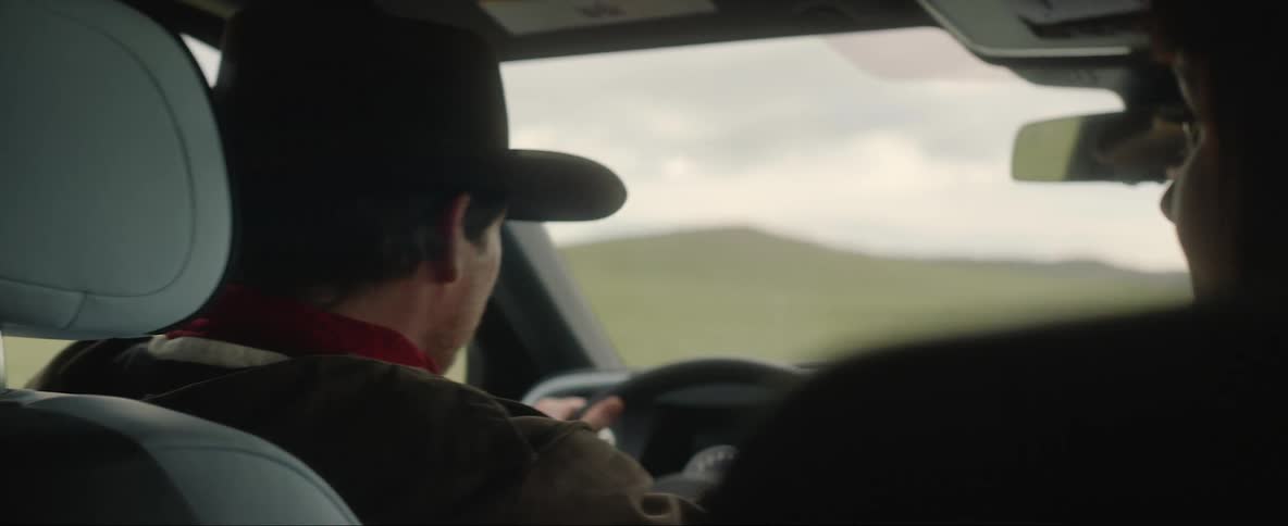 Man wearing cowboy hat drives car across plain briefly turning to smile at female passenger