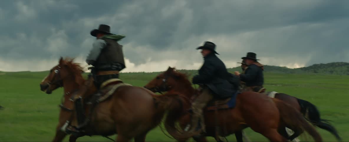 Four men wearing cowboy hats are riding horses across the plain under a cloudy sky