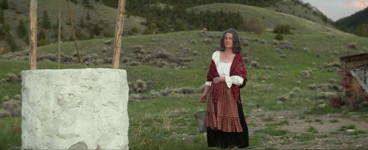 Woman wearing shawl walks from house to well with mountains and hills in the background