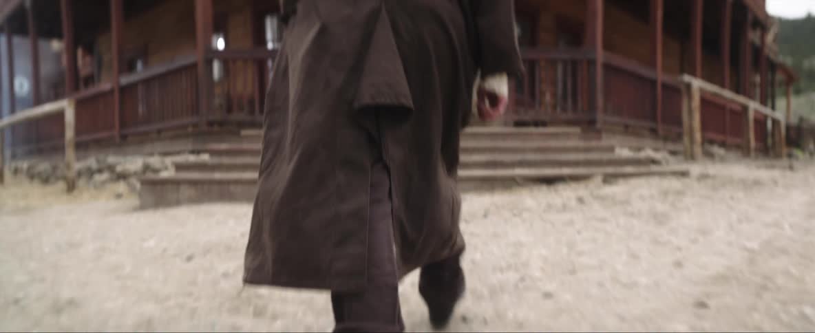 A man wearing a trench coat and a cowboy hat walks into a saloon