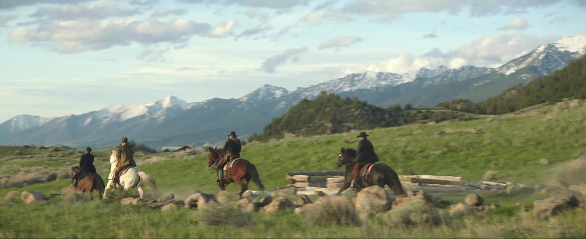 Horsemen wearing western wear ride along dirt road through valley near forested mountain