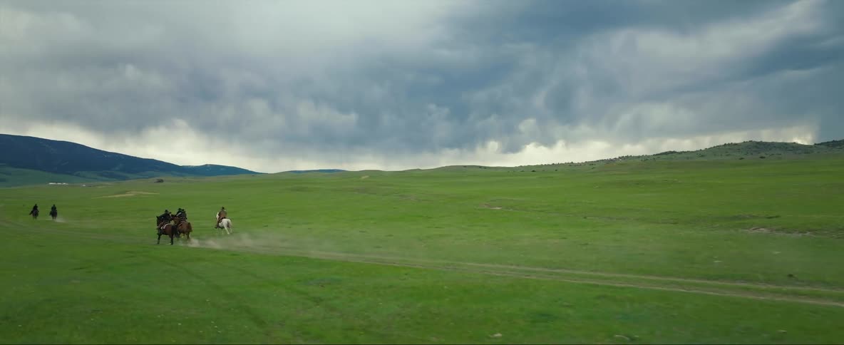Cowboys riding of on horses across a field under storm clouds